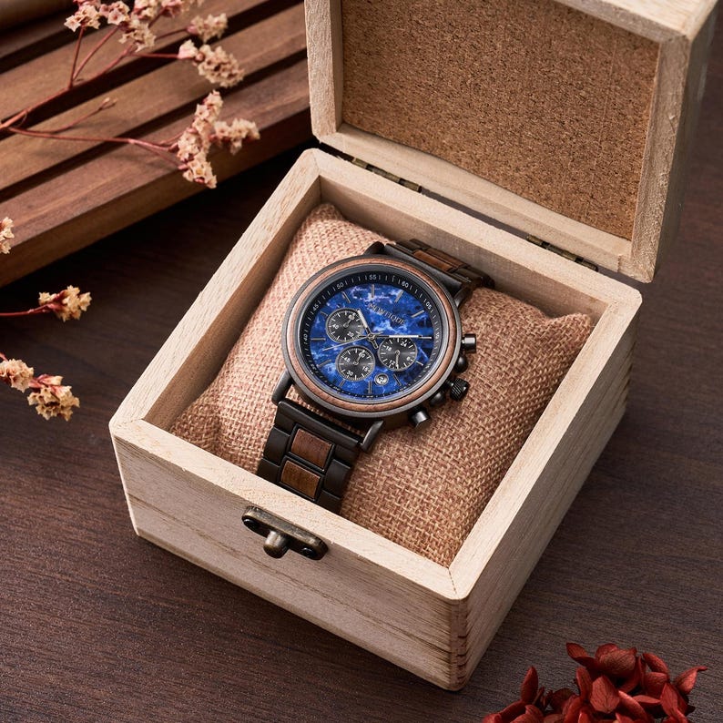 May include: A wooden wristwatch with a blue face and a brown and black band is displayed inside a wooden box. The watch has three sub-dials and a brown bezel. The box is lined with a tan fabric and has a small clasp.