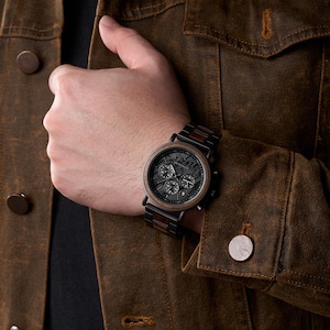 May include: A black and brown wooden watch with a black face and three sub-dials. The watch has a wooden bezel and a wooden and black metal band. The watch is on a person's wrist, and the person is wearing a brown jacket.