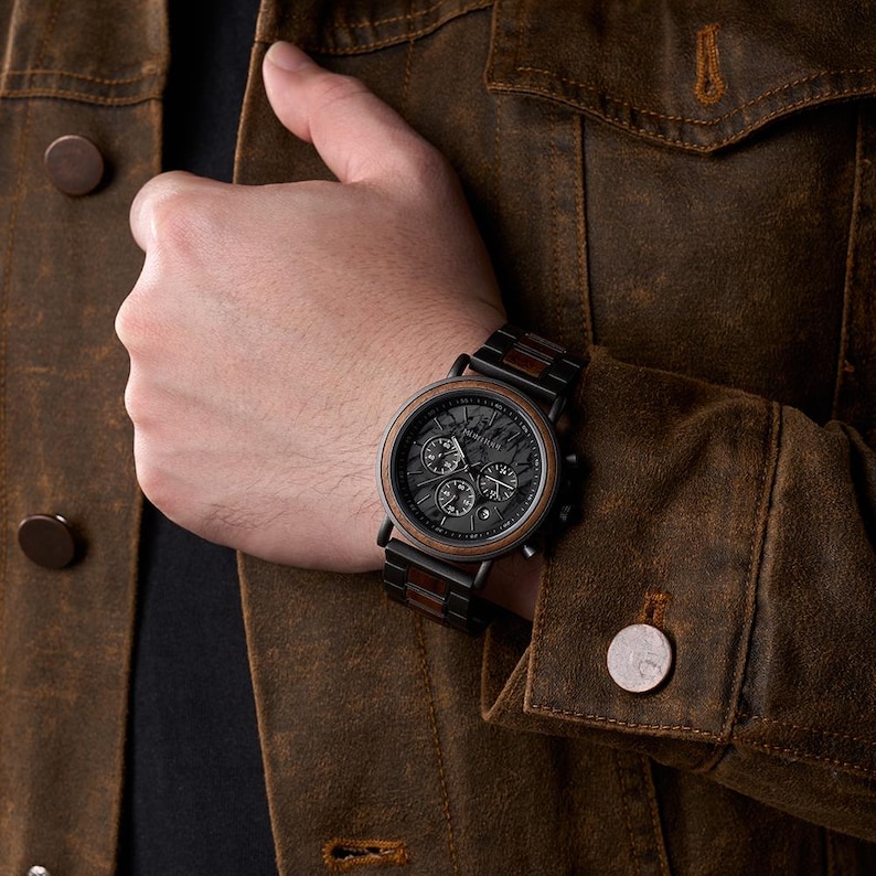 May include: A black and brown wooden watch with a black face and three sub-dials. The watch has a wooden bezel and a wooden and black metal band. The watch is on a person's wrist, and the person is wearing a brown jacket.