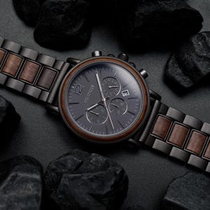 K&ouml;nnte beinhalten: Eine Armbanduhr mit einem Gliederarmband aus dunkelgrauem Metall und braunem Holz. Das runde Zifferblatt ist dunkelgrau mit dem Text MONTIQUE, einer gro&szlig;en 12, silbernen Zeigern, drei Hilfszifferbl&auml;ttern und einem Datumsfenster.