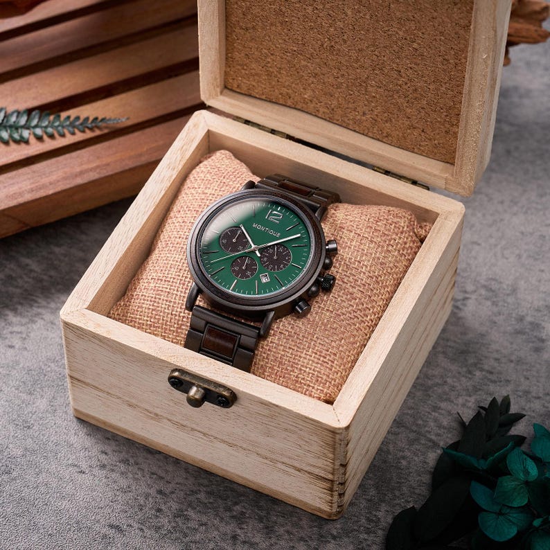 Puede incluir: Un reloj de madera con esfera verde y detalles negros, presentado en una caja de madera clara. El reloj lleva la marca "Montique" y muestra la fecha. La caja tiene un coj&iacute;n de tela de saco. La correa del reloj es marr&oacute;n oscuro.