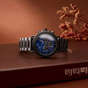 May include: A black wristwatch with a blue face and three sub-dials. The watch has a black metal band and a round black case. The watch is displayed on a brown surface with a blurred background.