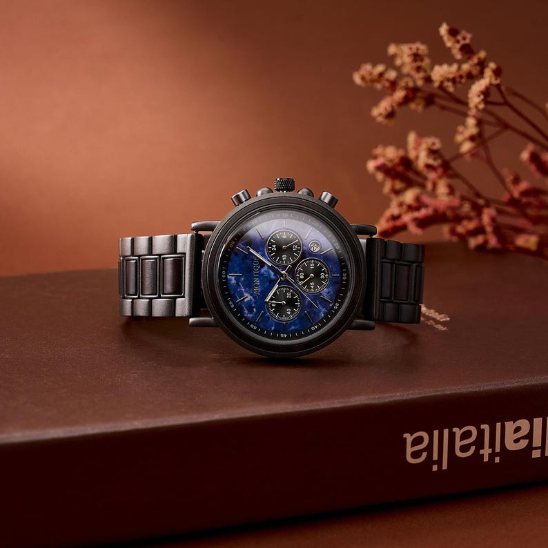 May include: A black wristwatch with a blue face and three sub-dials. The watch has a black metal band and a round black case. The watch is displayed on a brown surface with a blurred background.