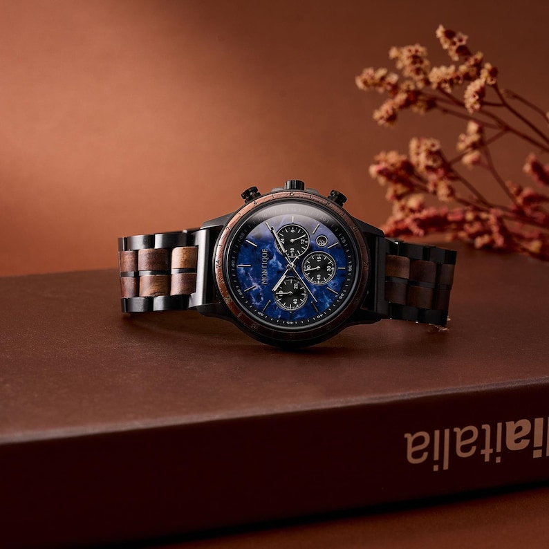 May include: A black and wood-toned wristwatch with a blue face and three sub-dials. The watch has a wooden band with alternating light and dark wood segments. The watch is displayed on a brown surface with a blurred background.