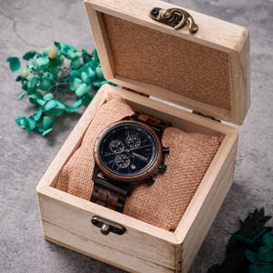 Op de afbeelding: Een houten horloge met een donkerblauwe wijzerplaat en bruine houten accenten, gepresenteerd in een lichtgekleurde houten doos met een scharnierend deksel. Het horloge heeft meerdere sub-wijzerplaten en de merknaam "MONTIQUE" is zichtbaar. De doos is gevoerd met een bruinachtige stof.