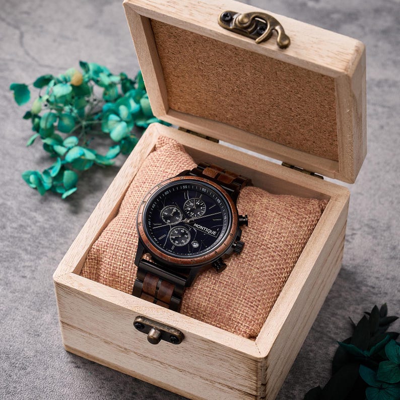 Op de afbeelding: Een houten horloge met een donkerblauwe wijzerplaat en bruine houten accenten, gepresenteerd in een lichtgekleurde houten doos met een scharnierend deksel. Het horloge heeft meerdere sub-wijzerplaten en de merknaam "MONTIQUE" is zichtbaar. De doos is gevoerd met een bruinachtige stof.