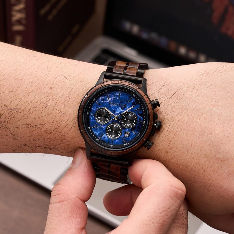 May include: A wristwatch with a dark wood grain band and a black case. The watch face is blue with three smaller dials and the brand name "Muntere". The watch is on a wrist.