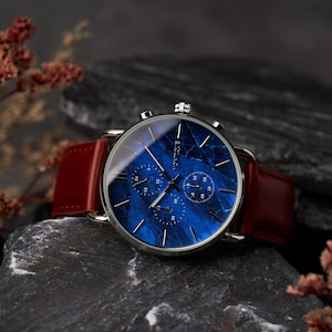 May include: A silver-toned wristwatch with a blue marble-patterned face and three sub-dials. The watch has a brown leather band. The word "Vontique" is printed on the watch face. The watch is displayed on a dark gray stone surface.