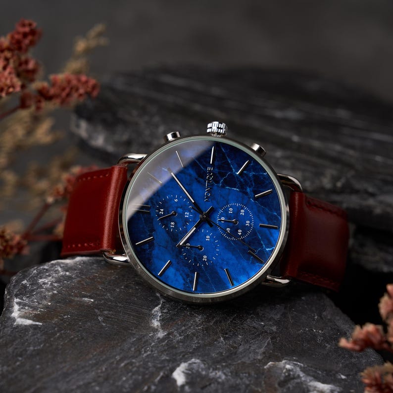 May include: A silver-toned wristwatch with a blue marble-patterned face and three sub-dials. The watch has a brown leather band. The word "Vontique" is printed on the watch face. The watch is displayed on a dark gray stone surface.