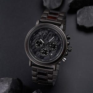 May include: A black wristwatch with a dark marble-like face and three sub-dials. The watch has a black metal band and a wooden accent. The watch is displayed on a dark surface with dark rocks.