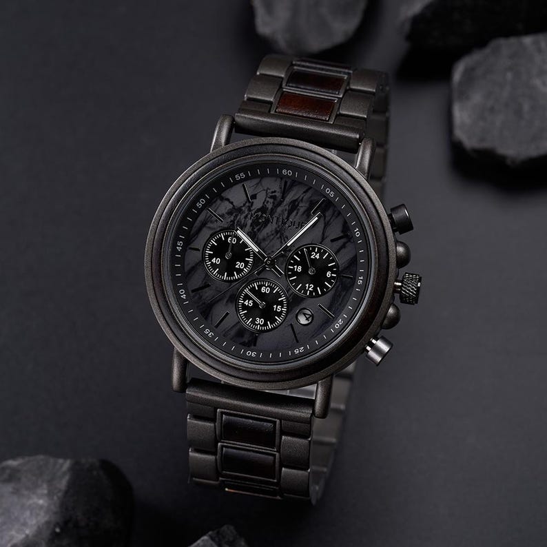 May include: A black wristwatch with a dark marble-like face and three sub-dials. The watch has a black metal band and a wooden accent. The watch is displayed on a dark surface with dark rocks.