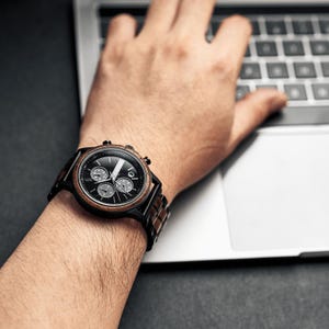Op de afbeelding: Een zwart en bruin polshorloge met een zwarte wijzerplaat en drie sub-wijzerplaten wordt getoond op een pols. Het horloge heeft een houten band en een zwarte bezel. Het horloge wordt gedragen terwijl een persoon een laptop gebruikt.
