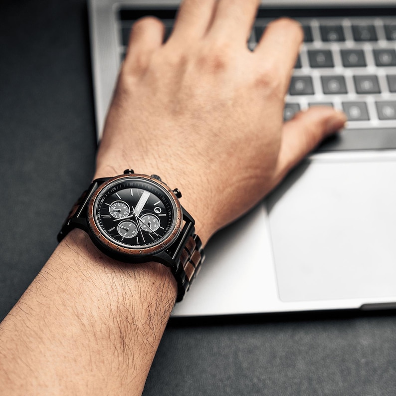 Op de afbeelding: Een zwart en bruin polshorloge met een zwarte wijzerplaat en drie sub-wijzerplaten wordt getoond op een pols. Het horloge heeft een houten band en een zwarte bezel. Het horloge wordt gedragen terwijl een persoon een laptop gebruikt.