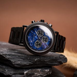 May include: A black wristwatch with a blue face and black band. The watch face has three smaller dials and the brand name "MORTIQUE". The watch is displayed on a dark gray rock against a brown background.