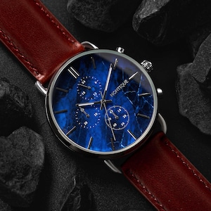 May include: A wristwatch with a blue marble-patterned face and silver casing. The watch features three sub-dials and the brand name "NONTIQUE". It has a brown leather strap. The watch is displayed on a dark surface with black rocks.