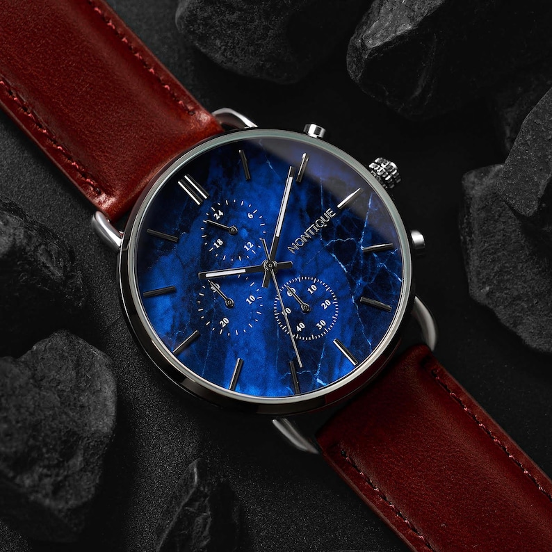 May include: A wristwatch with a blue marble-patterned face and silver casing. The watch features three sub-dials and the brand name "NONTIQUE". It has a brown leather strap. The watch is displayed on a dark surface with black rocks.