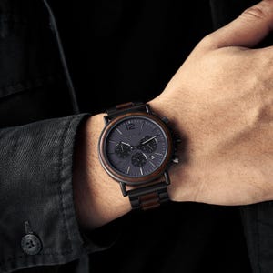 K&ouml;nnte beinhalten: Eine Chronographen-Armbanduhr mit dunkelgrauem Zifferblatt, einer markanten Zahl 12, drei Hilfszifferbl&auml;ttern und einer Datumsanzeige. Das Geh&auml;use und das Armband bestehen aus einer Kombination von mattschwarzem Metall und dunkelbraunen Holzakzenten.