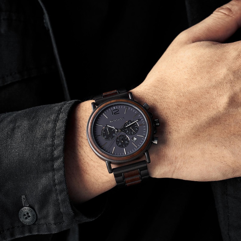K&ouml;nnte beinhalten: Eine Chronographen-Armbanduhr mit dunkelgrauem Zifferblatt, einer markanten Zahl 12, drei Hilfszifferbl&auml;ttern und einer Datumsanzeige. Das Geh&auml;use und das Armband bestehen aus einer Kombination von mattschwarzem Metall und dunkelbraunen Holzakzenten.