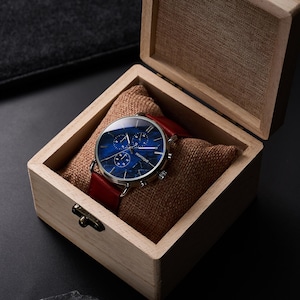 May include: A wristwatch with a blue face and red leather band rests in a wooden box lined with burlap. The watch face has a unique marbled design. The box is open, revealing the watch and its presentation.
