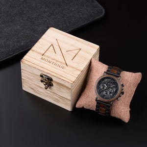 May include: A wooden watch with a black face and brown accents rests on a small pillow. The watch has three sub-dials and a dark metal band. A wooden box with the word "MONTIQUE" is next to the watch.
