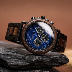 May include: A wristwatch with a blue face and wooden accents. The watch features a black bezel and a wooden band. The watch face has three sub-dials and the brand name "MONTIQUE". The watch is resting on a dark gray stone.