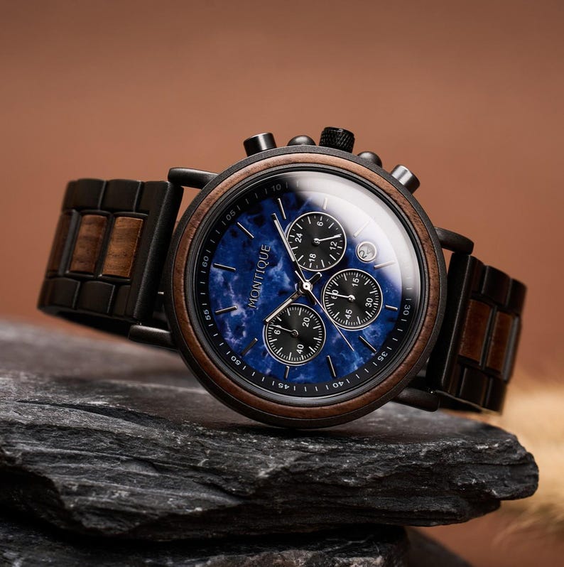 May include: A wristwatch with a blue face and wooden accents. The watch features a black bezel and a wooden band. The watch face has three sub-dials and the brand name "MONTIQUE". The watch is resting on a dark gray stone.