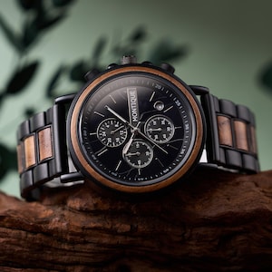 Może przedstawiać: Czarny zegarek chronograf Montique z drewnianą ramką i paskiem. Tarcza zegarka jest czarna z trzema tarczami pomocniczymi i okienkiem daty. Pasek posiada drewniane akcenty. Zegarek jest prezentowany na kawałku drewna.