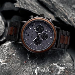K&ouml;nnte beinhalten: Eine Chronographen-Armbanduhr mit dunkelgrauem Zifferblatt und der Aufschrift Montique, einer braunen Holzl&uuml;nette sowie einem Armband aus abwechselnden schwarzen Metall- und braunen Holzgliedern. Die Uhr verf&uuml;gt &uuml;ber drei Hilfszifferbl&auml;tter und ein Datumsfenster.