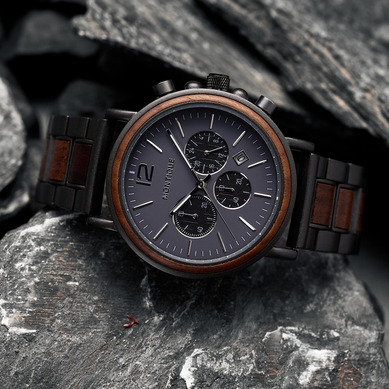 K&ouml;nnte beinhalten: Eine Chronographen-Armbanduhr mit dunkelgrauem Zifferblatt und der Aufschrift Montique, einer braunen Holzl&uuml;nette sowie einem Armband aus abwechselnden schwarzen Metall- und braunen Holzgliedern. Die Uhr verf&uuml;gt &uuml;ber drei Hilfszifferbl&auml;tter und ein Datumsfenster.