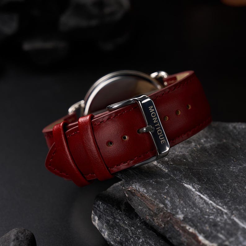 May include: Close-up of a wristwatch with a red leather band. The watch has a silver buckle with the word "Montique" engraved on it. The watch is set against a dark background.