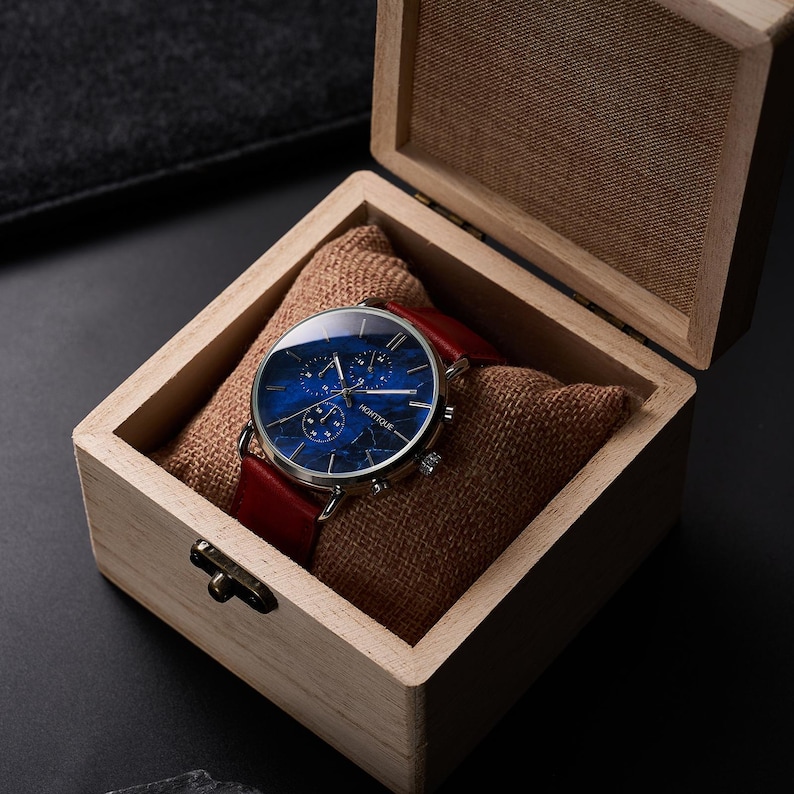May include: A wristwatch with a blue face and red leather band rests in a wooden box lined with burlap. The watch face has a marbled design and silver accents. The box is open, revealing the watch and its presentation.