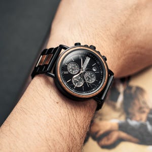 May include: A black wristwatch with a wooden bezel and band accents. The watch face is black with three sub-dials and the brand name "MONTIQUE". The watch is on a wrist.