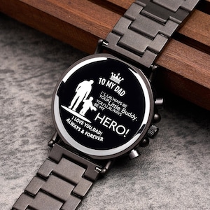 Puede incluir: Reloj de pulsera negro con una correa de metal gris oscuro. La esfera del reloj presenta el texto "TO MY DAD" y "HERO!" junto con una silueta de padre e hijo. El reloj tambi&eacute;n incluye las frases "I LOVE YOU, DAD! ALWAYS & FOREVER."