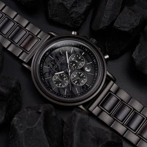 May include: A black Montique watch with a marble-patterned face and three sub-dials. The watch has a black metal band with wooden accents and is surrounded by dark rocks. The watch face displays the time and date.