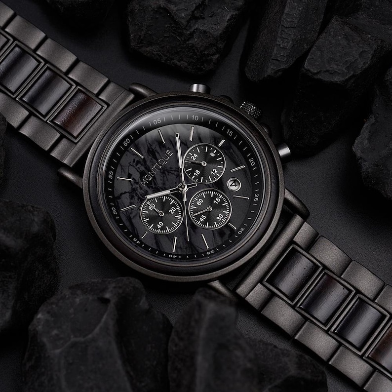 May include: A black Montique watch with a marble-patterned face and three sub-dials. The watch has a black metal band with wooden accents and is surrounded by dark rocks. The watch face displays the time and date.