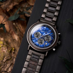 May include: A wooden wristwatch with a dark gray band and a blue face. The watch face has three smaller dials and the word "Montique" is printed on the face. The watch is displayed on a black surface.