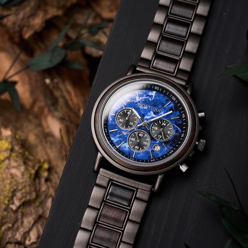 May include: A wooden wristwatch with a dark gray band and a blue face. The watch face has three smaller dials and the word "Montique" is printed on the face. The watch is displayed on a black surface.