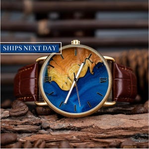 Peut inclure: Une montre-bracelet dorée avec un bracelet en cuir marron. Le cadran de la montre présente un design bleu et boisé avec des chiffres romains pour les heures.
