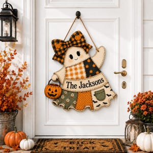 May include: A hanging door sign shaped like a ghost wearing a large orange and black plaid bow. The ghost features a patchwork-style dress with pumpkin, floral, and bat patterns, holding a small jack-o'-lantern bucket with the text The Jacksons.