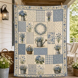 May include: A rectangular quilt featuring a patchwork design of blue and cream floral patterns, bouquets in pitchers, watering cans, and baskets. The quilt is bordered by a blue floral vine print and hangs from a wooden rod on a porch.