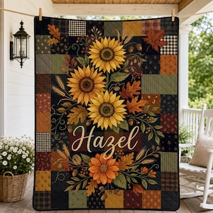 May include: A quilted blanket featuring a patchwork design of dark brown, green, and black squares. The center displays a vibrant illustration of yellow sunflowers, orange flowers, autumn leaves, and acorns with the name Hazel in white script.