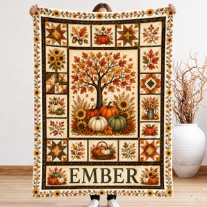 May include: A rectangular throw blanket featuring a central illustration of a tree with autumn leaves, pumpkins, and sunflowers. The border contains smaller panels of geometric quilt patterns, foliage, and the word EMBER in large letters.