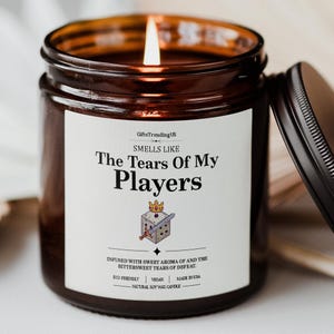 May include: A lit soy wax candle in a brown glass jar with a black lid. The label reads "Smells Like The Tears Of My Players" with a dice and sword graphic. The candle is eco-friendly, vegan, and made in the USA.