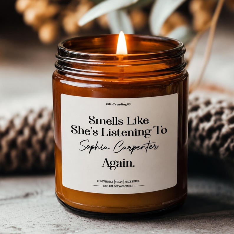 May include: An amber glass candle with a lit wick and a white label. The label says "Smells Like She's Listening To Sophia Carpenter Again." The candle is eco-friendly, vegan, and made in the USA. It is a natural soy wax candle.