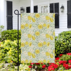 May include: A garden flag featuring a repeating pattern of yellow sunflowers, lemons, and white blossoms on a light green background. The flag is displayed outdoors, adding a touch of floral charm to the yard.