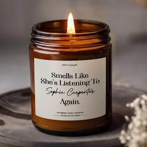 May include: A burning candle in a brown glass jar. The candle has a white label with the text "Smells Like She's Listening To Sophia Carpenter Again." The candle is made from natural soy wax and is eco-friendly.