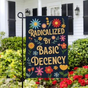 May include: A navy blue garden flag with the words "RADICALIZED BY BASIC DECENCY" in yellow script, surrounded by colorful floral illustrations. The flag is displayed outdoors, adding a touch of whimsy to the garden.