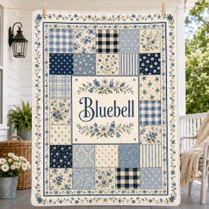 May include: A rectangular patchwork quilt featuring a cream-colored center panel with the text Bluebell in navy blue script, surrounded by floral and botanical illustrations. The quilt is composed of various blue and white patterned squares.