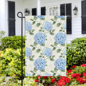 May include: A garden flag featuring a repeating pattern of blue hydrangea flowers with green leaves and stems on a cream background. The text "F*ck Trump" is printed on the stems. The flag is displayed outdoors.