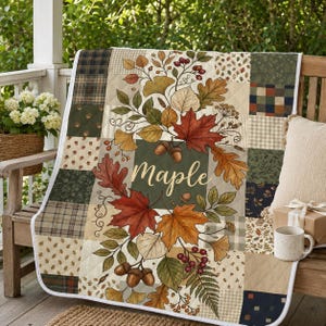 May include: A quilted throw blanket featuring a patchwork design of plaid and autumn-themed patterns. The center displays a botanical illustration of maple leaves, acorns, and berries with the word Maple written in a cursive script font.
